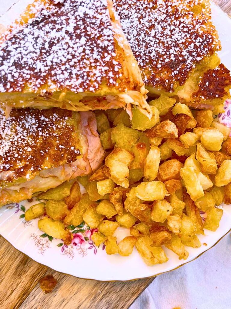 An overhead view of a sliced Monte Cristo sandwich dusted with powdered sugar. There are crispy potato cubes on the side of the sandwich.