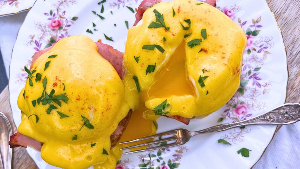 An overhead view of two eggs Benedict on a pink floral plate. Once eggs has been split to show the runny yolk inside.