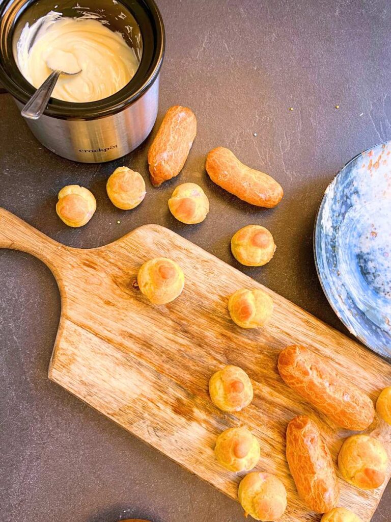 Profiteroles on a wooden cutting board. There is melted white chocolate off to the side.