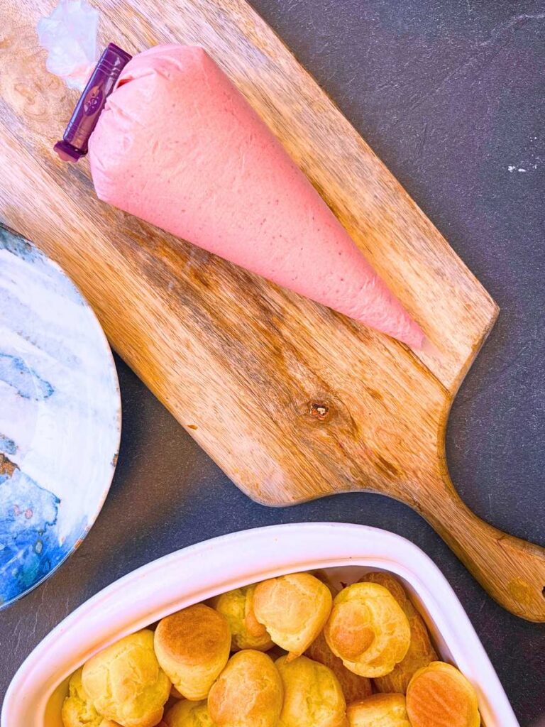 A piping bag with strawberry cream. There are profiteroles and a blue bowl.