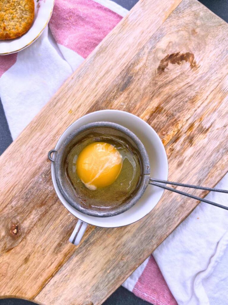 A cracked egg in a small metal strainer overtop of a teacup.
