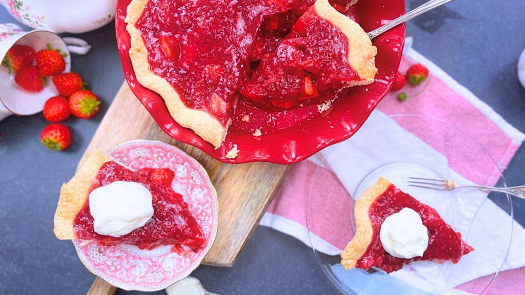 A sliced strawberry pie in a red pie plate. there are two slices in front with whipped cream on top.