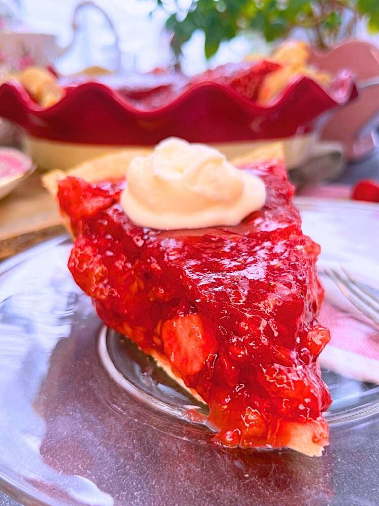 A slice of strawberry pie with whipped cream on top. There is more pie in a red pie plate in the background.