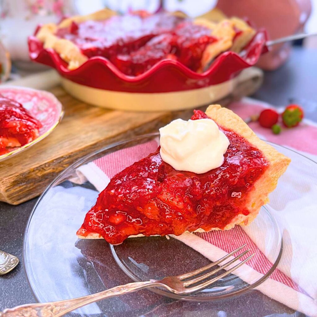A side view of a slice of strawberry pie. There is more of the pie in the background in a red pie plate.