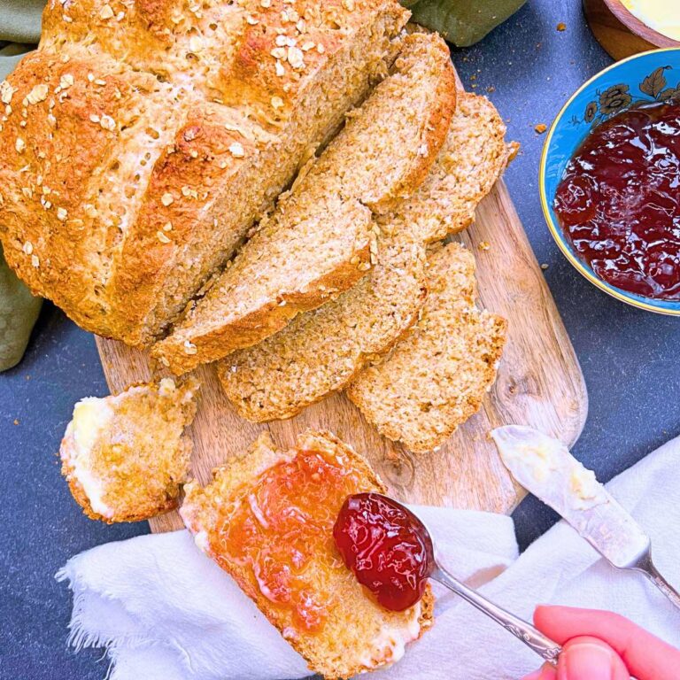 An overhead view of an Irish soda bread that is partially sliced. A woman is spooning jam on one of the slices.