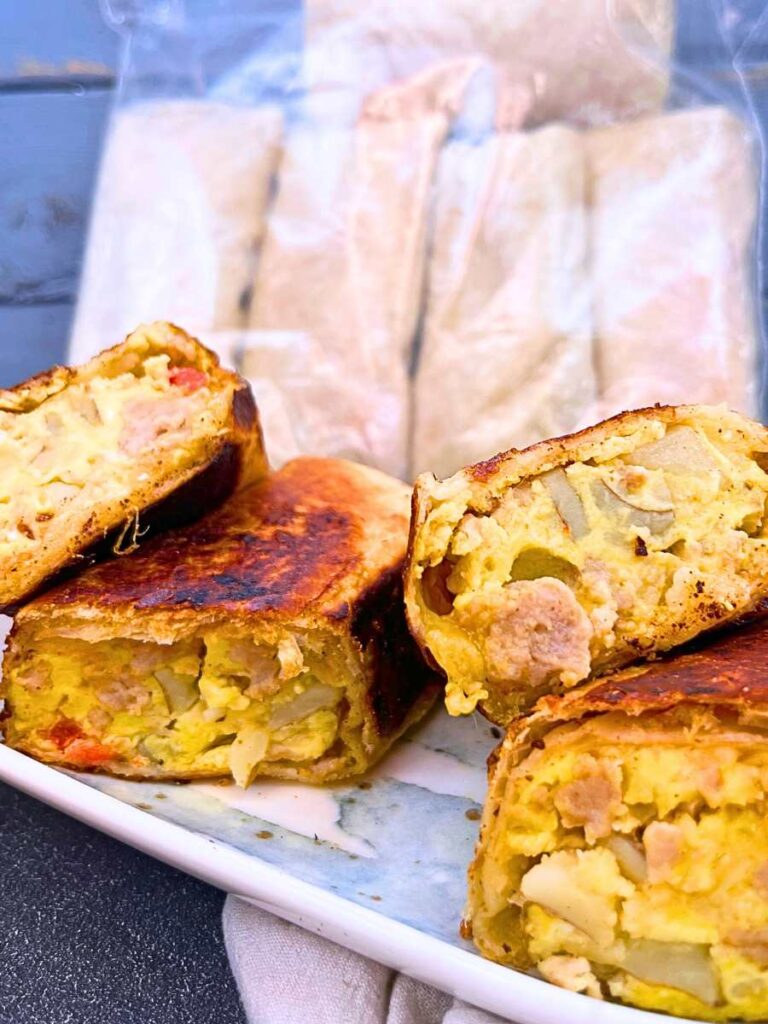 Two breakfast burritos that have been cut in half to show the inside filling. There are more in the background in a freezer bag.