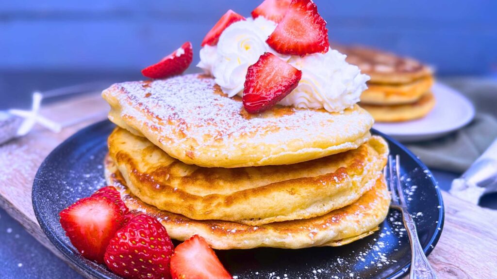 A side view of a stack of three buttermilk pancakes. They are garnished with whipped cream and strawberries