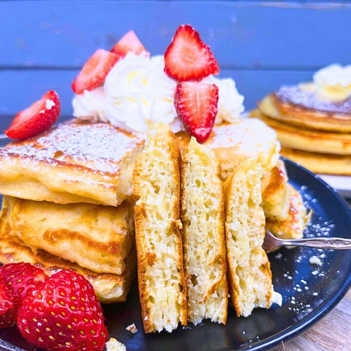A stack of buttermilk pancakes with one slice removed and turned on it's side. They are garnished with strawberries and whipped cream.