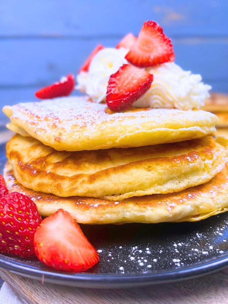 A side view of a stack of pancakes on a black plate. They are topped with whipped cream and sliced strawberries.