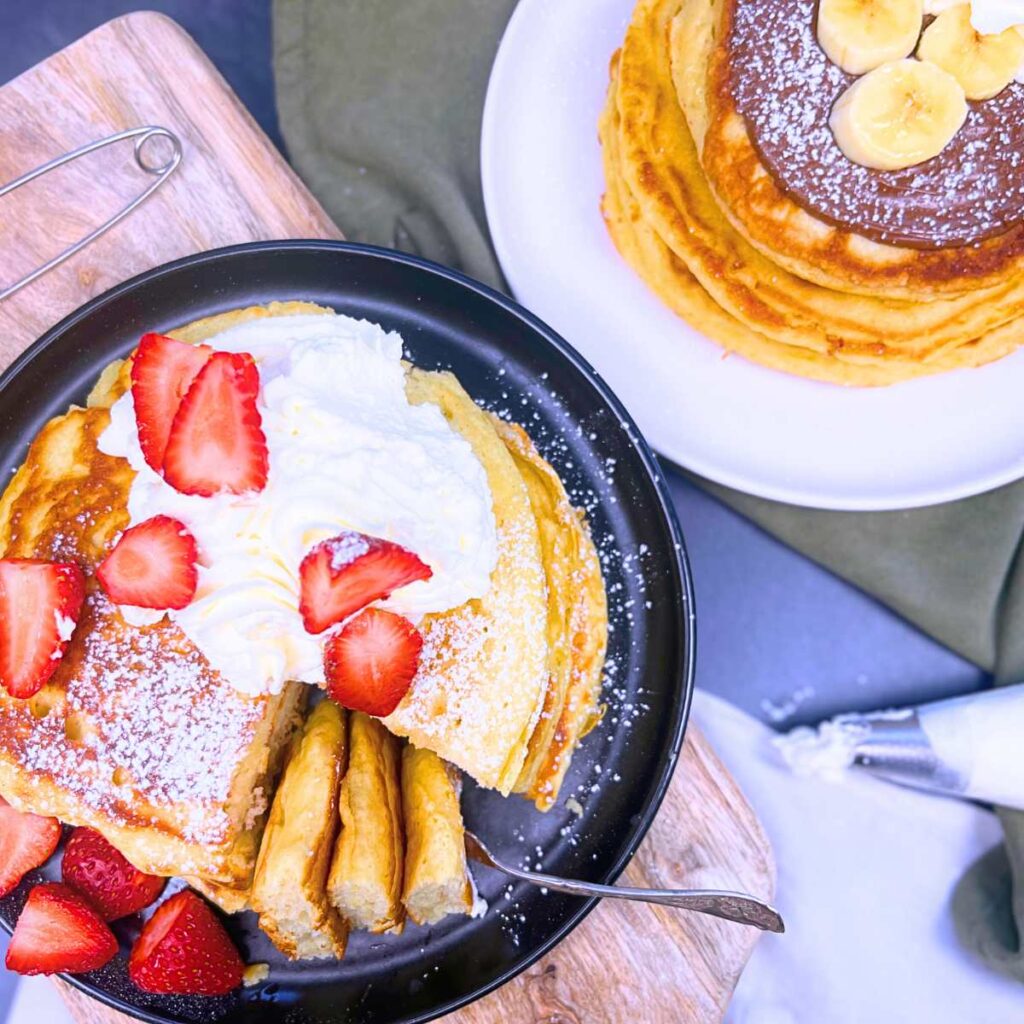 Two plates of buttermilk pancakes. One is topped with strawberries and whipped cream. The other is topped with Nutella and banana slices.