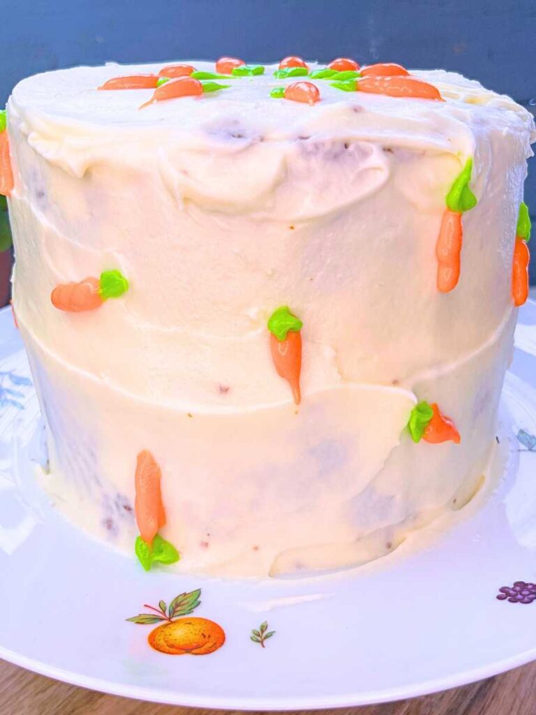 A cake on a cake stand covered with white icing. It is decorated with icing carrots.