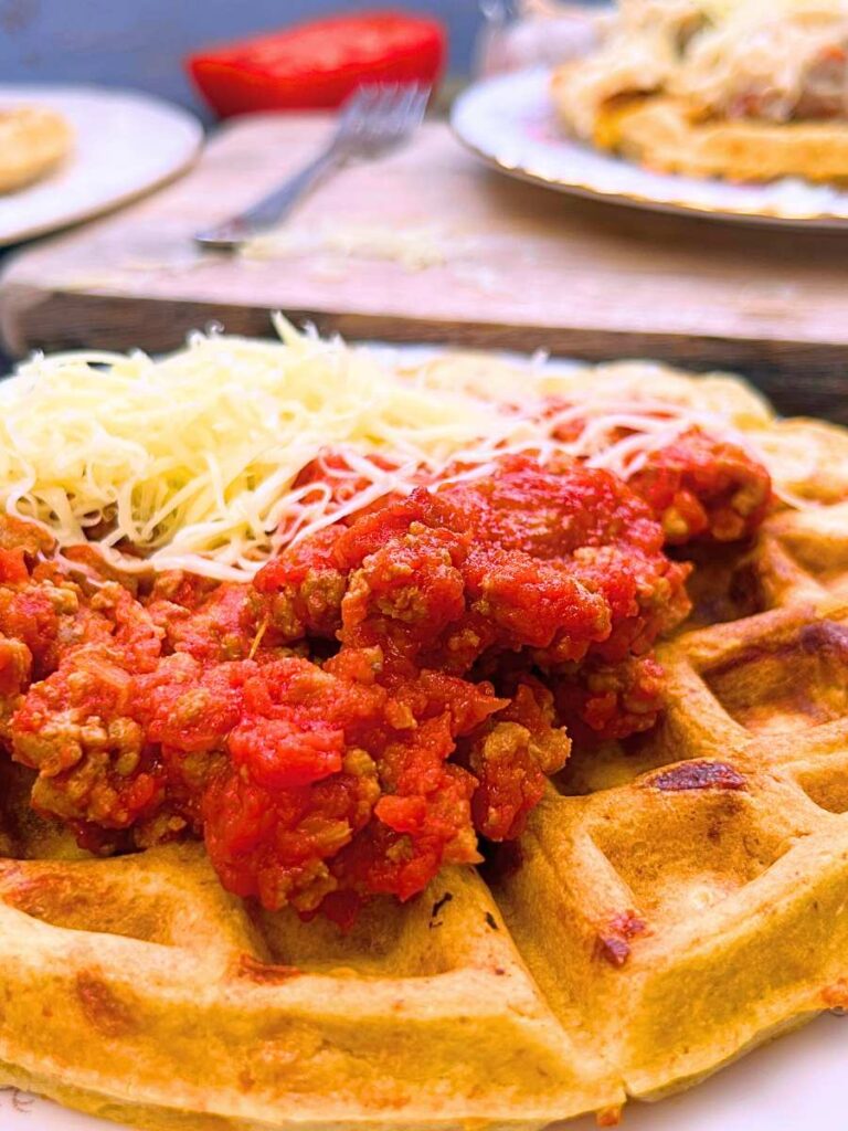 A side view of a cheese waffle with meat and tomato sauce on top.