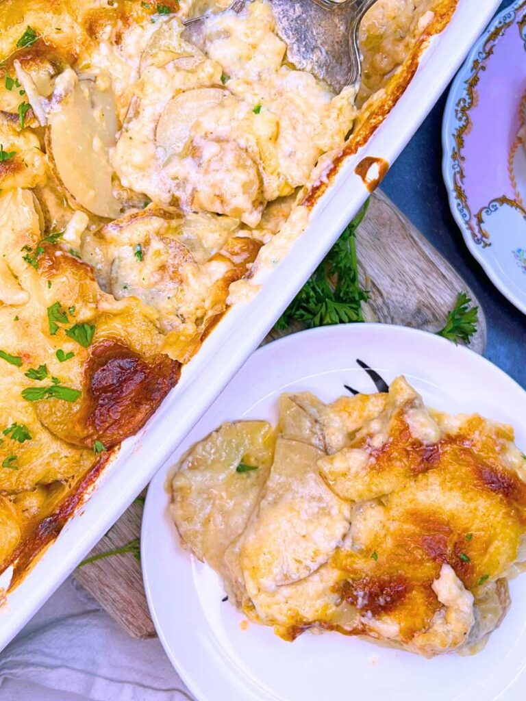 Scalloped potatoes in a white casserole dish. Threre is a serving of to the side on a white plate.