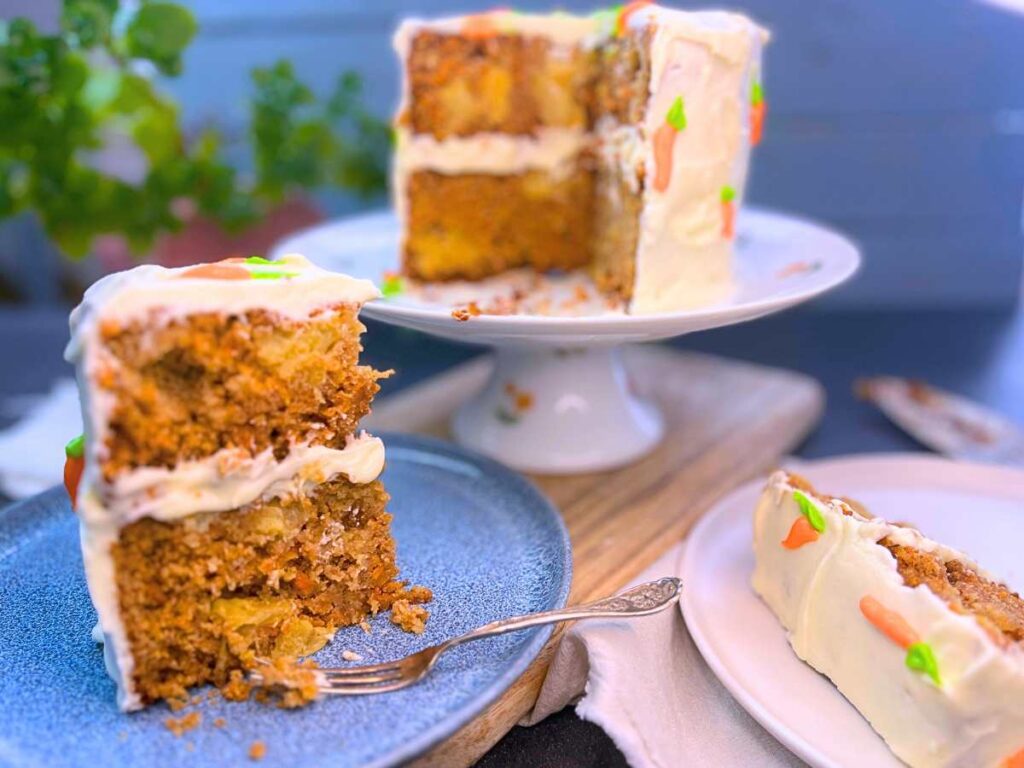 A carrot cake on a cake stand. There are two slices in front. One is on it's side. The cake is decorated with cream cheese with icing and icing carrots.
