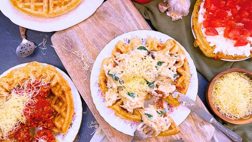 Three topped savoury waffles. Once has cream cheese and tomatoes, one has a cheesy chicken, tomato, and spinach mixture. The last is topped with a ground meat and tomato sauce.