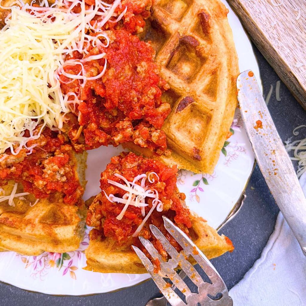 A woman is taking a slice from a savoury cheese waffle that has a tomato and meat sauce on top.