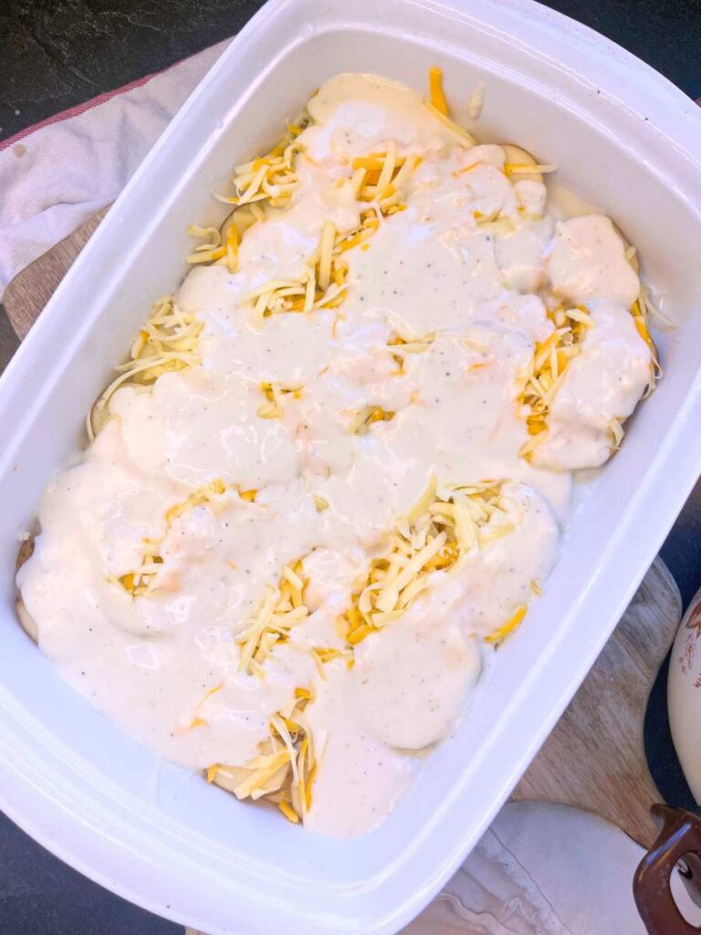 A white casserole dish white sliced potatoes, grated cheese, and béchamel sauce.