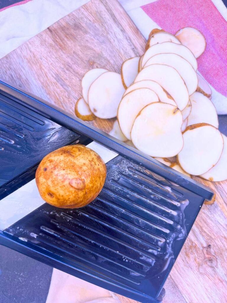A mandolin with slices of potatoes. There is half of a potato on top of the mandolin.