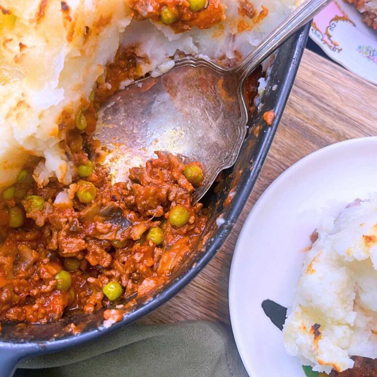 An overhead view of a shepherd's pie casserole with a serving spoon inside. There is a serving on a white plate off to the side.