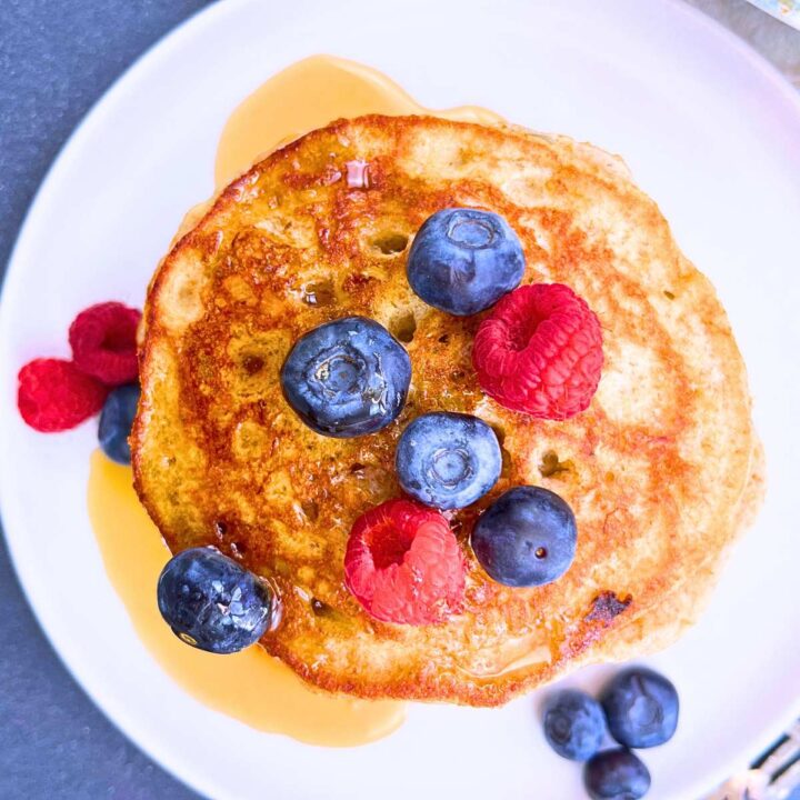An overhead view of a stack of pancakes with maple syrup and fresh berries on a white plate.