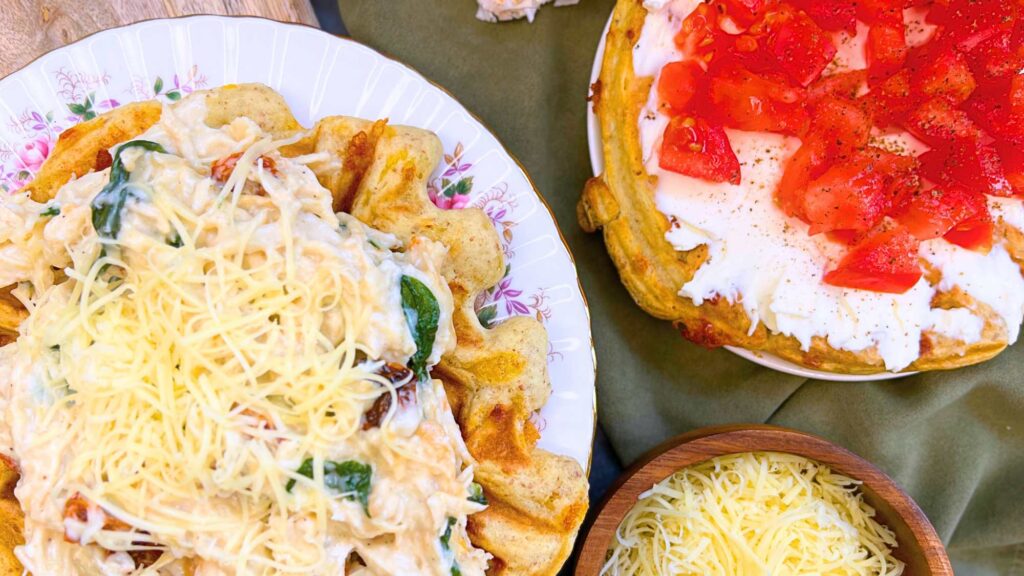 An overhead view of two sourdough savoury cheese waffles. Once is spread with cream cheese and tomatoes. The other is topped with a creamy chicken, tomato, and spinach mixture.