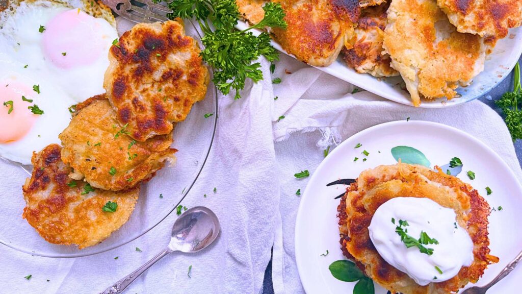 Potato pancakes on a plate topped with sour cream. There are more on a plate with over easy eggs and a platter of Irish boxty pancakes in the background.