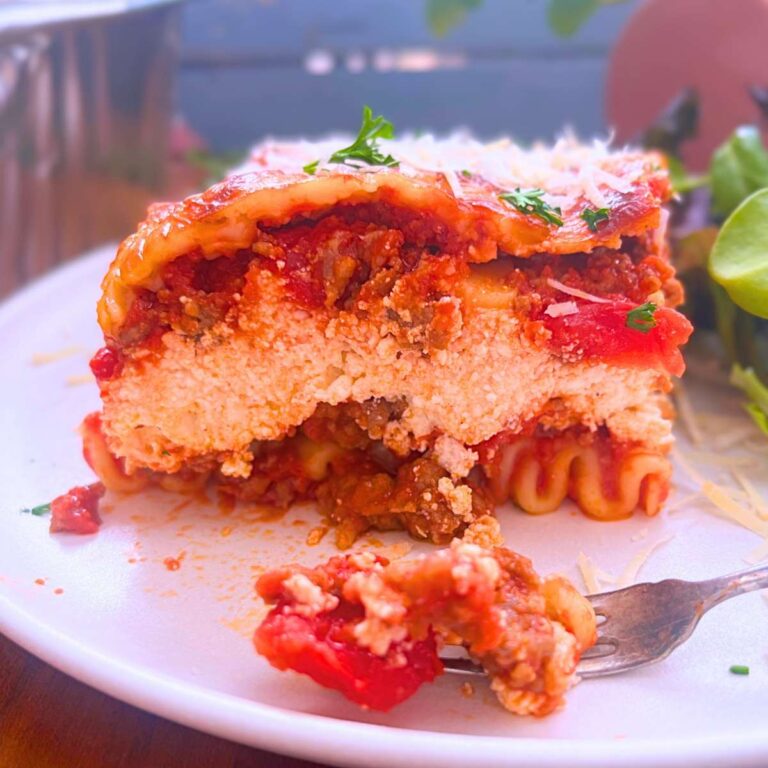 A side view of a baked lasagna, showing the layers inside. There is a forkful on front on a white plate.