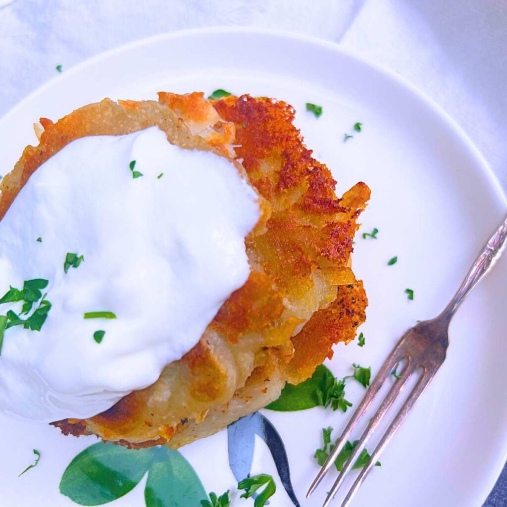 A white plate with a stack of potato pancakes. They are topped with sour cream.