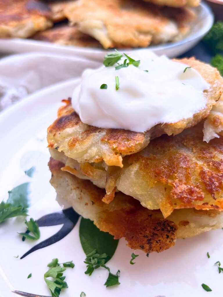 A side view of a stack of boxty potato pancakes on a white plate. They are topped with sour cream.