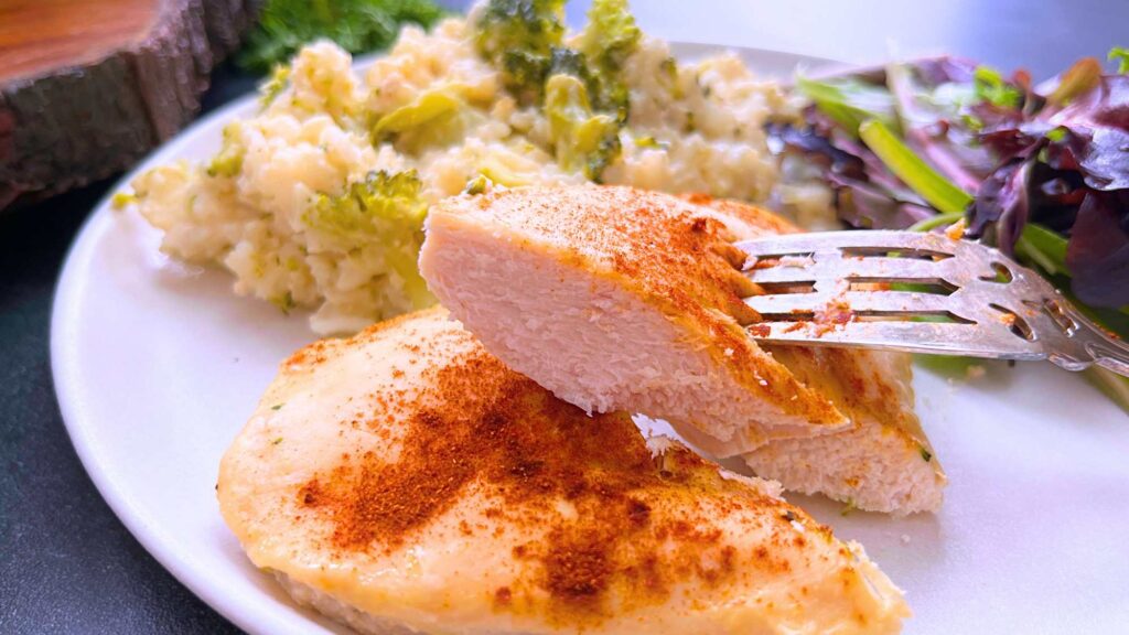 A woman is holding half of a cooked chicken breast on a fork. There is more chicken on the plate with rice and broccoli in the background.