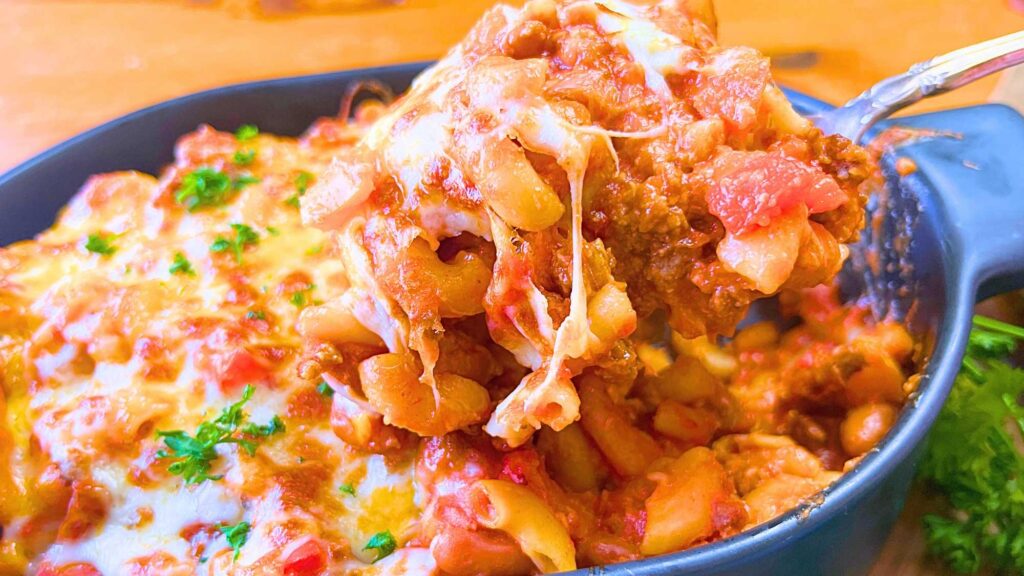 A woman is lifting a serving spoon with chili mac and cheese out of a black casserole dish.