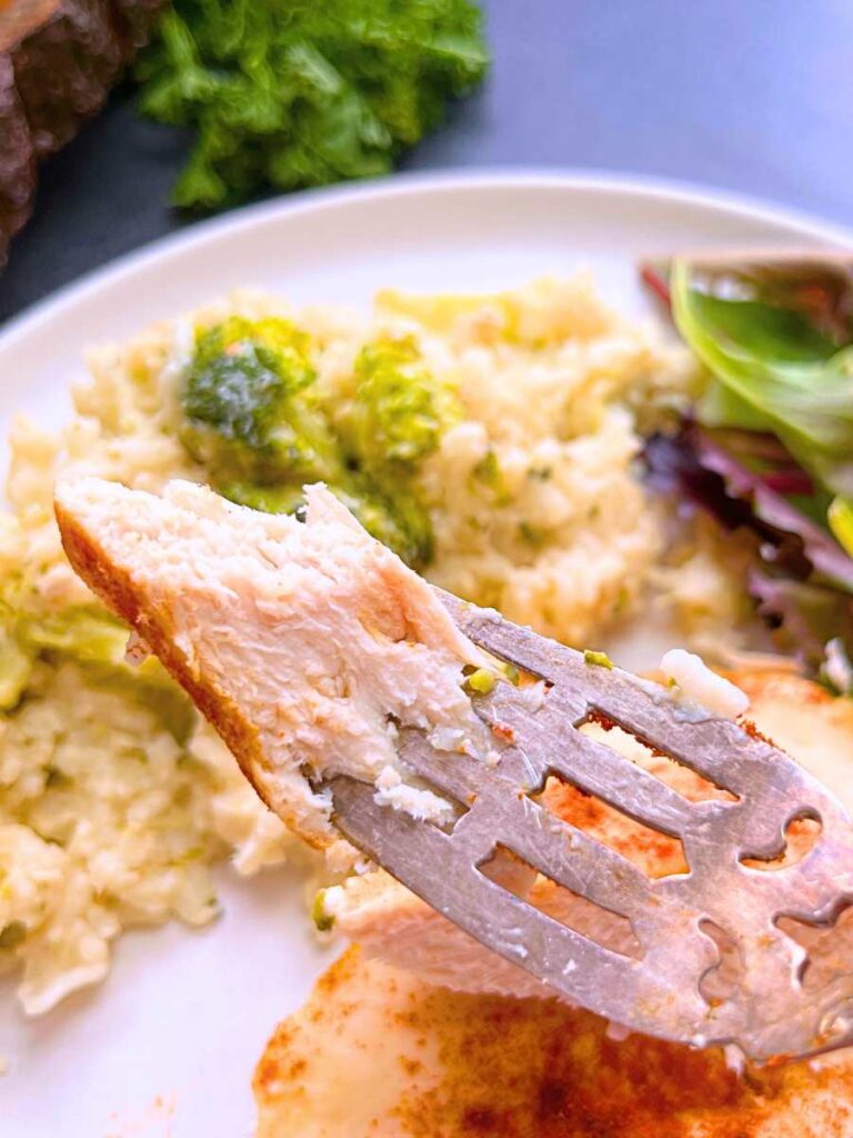 A woman is holding up a forkful of chicken breast. There is more chicken with a side serving of broccoli, rice, and a side salad in the background.
