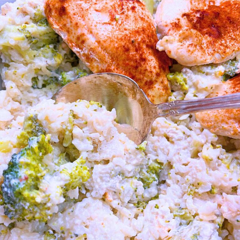 A woman is holding a large metal serving spoon in a casserole of rice, broccoli, and chicken breast garnished with paprika.