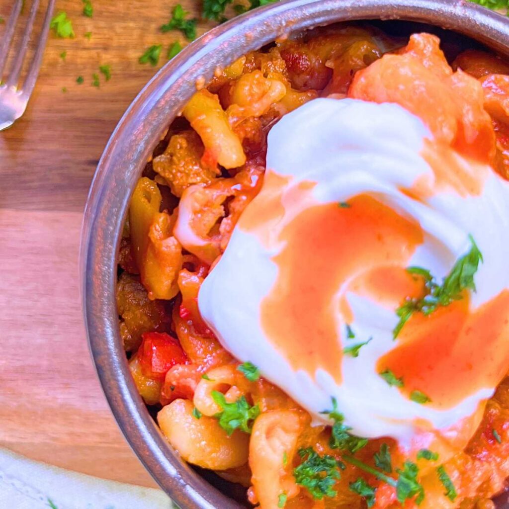 A brown bowl with a serving of chili mac and cheese topped with sour cream and hot sauce.