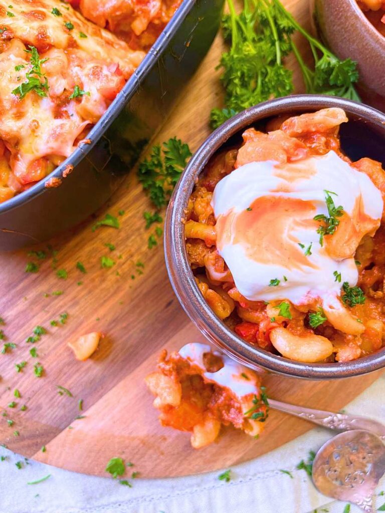 A brown bowl with a serving of chili mac and cheese topped with sour cream and hot sauce. There is more in a black casserole dish in the background.