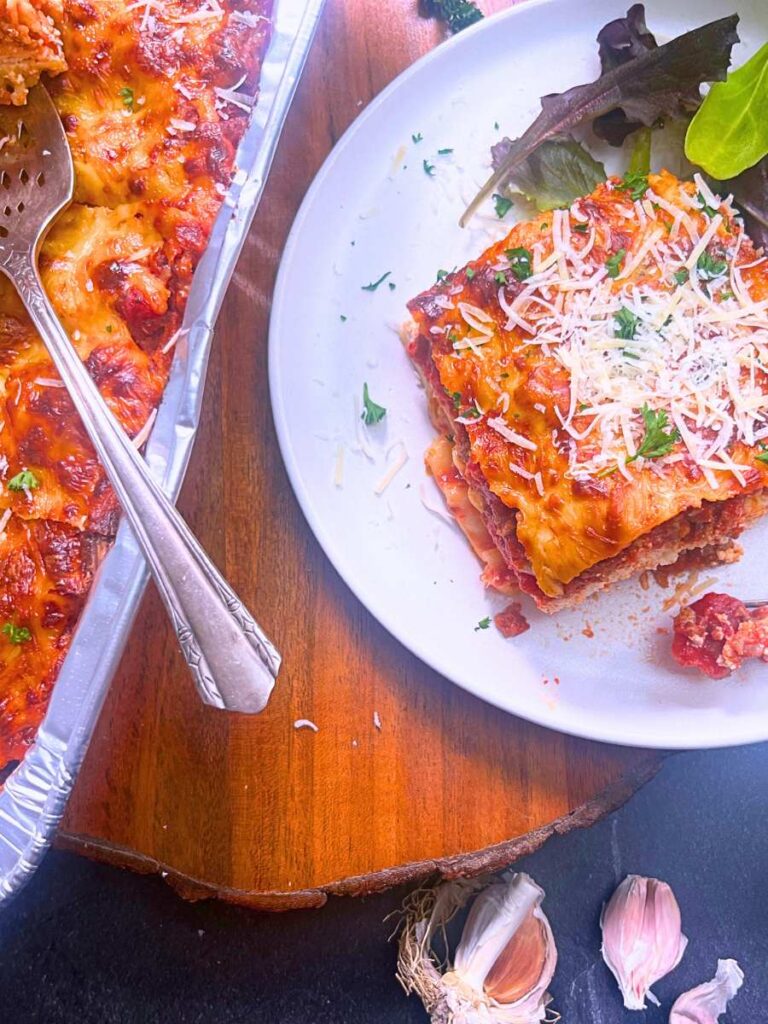 An over head view of a baked lasagna in an aluminum tin. There is a serving on a white plate off to the side. There is a salad on the side.