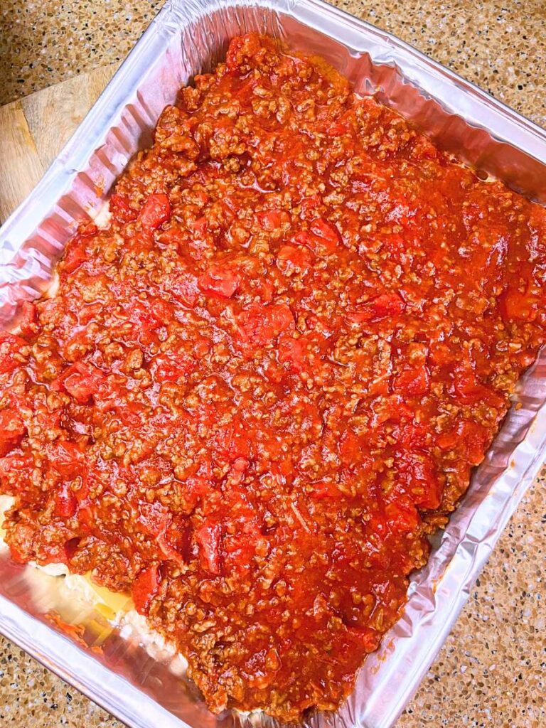 An aluminum casserole dish with an assembled lasagna with the meat and tomato sauce layer on top.