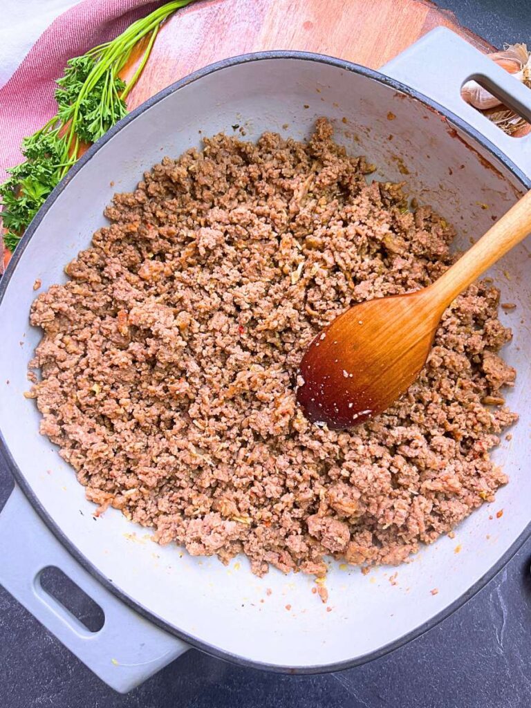 A grey Dutch oven with cooked ground beef and ground sausage. There is a wooden spoon inside.