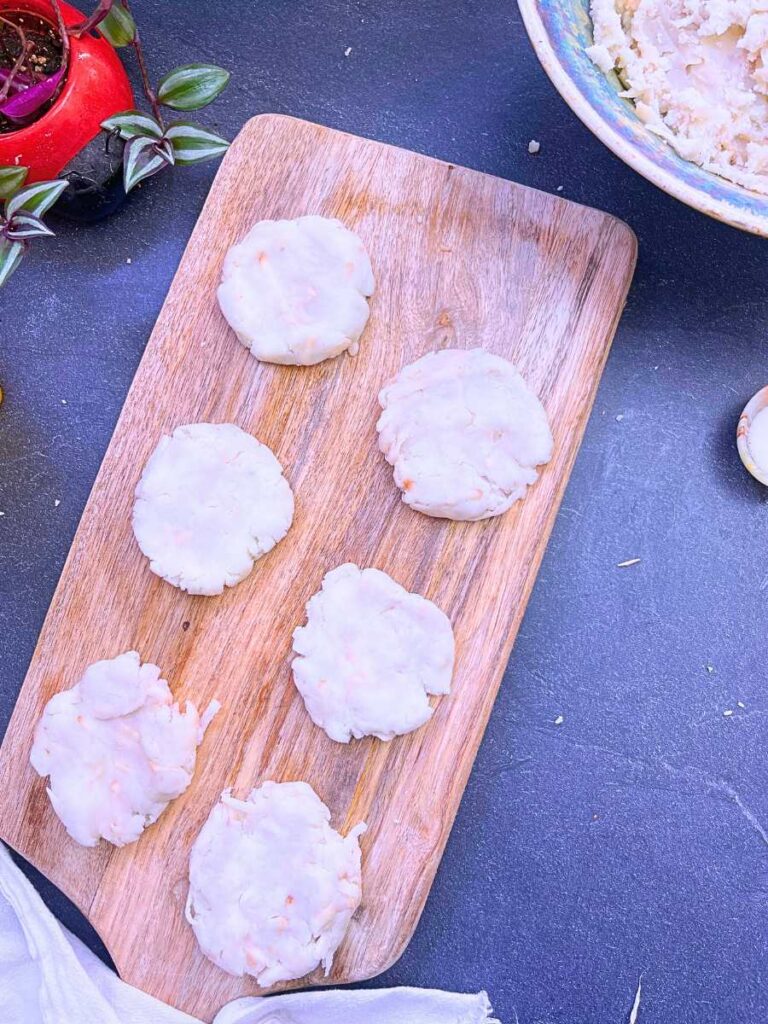 Six uncooked potato pancakes on a wooden cutting board.