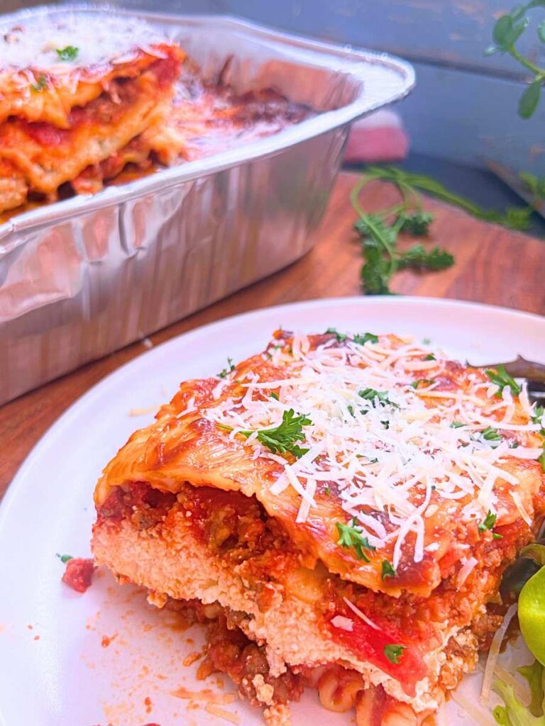 A serving of baked lasagna on a white plate with a salad on the side. There is more in the background in an aluminum pan.