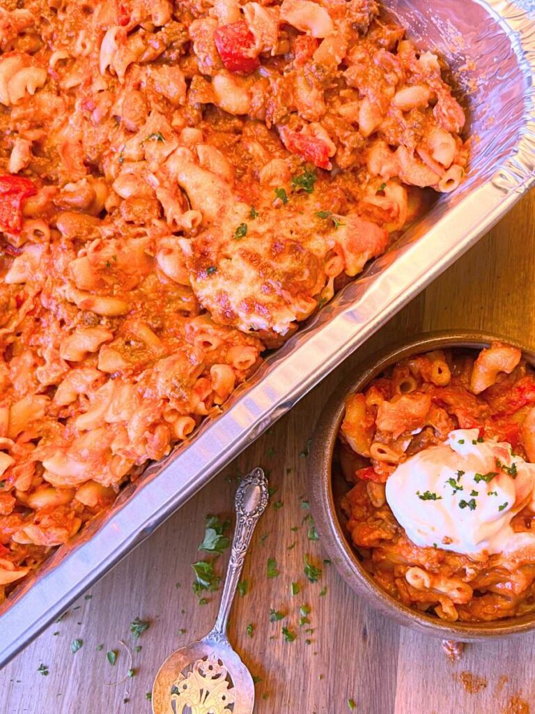 Chili mac and cheese in a aluminum tray. There is a serving in front topped with sour cream and hot sauce.