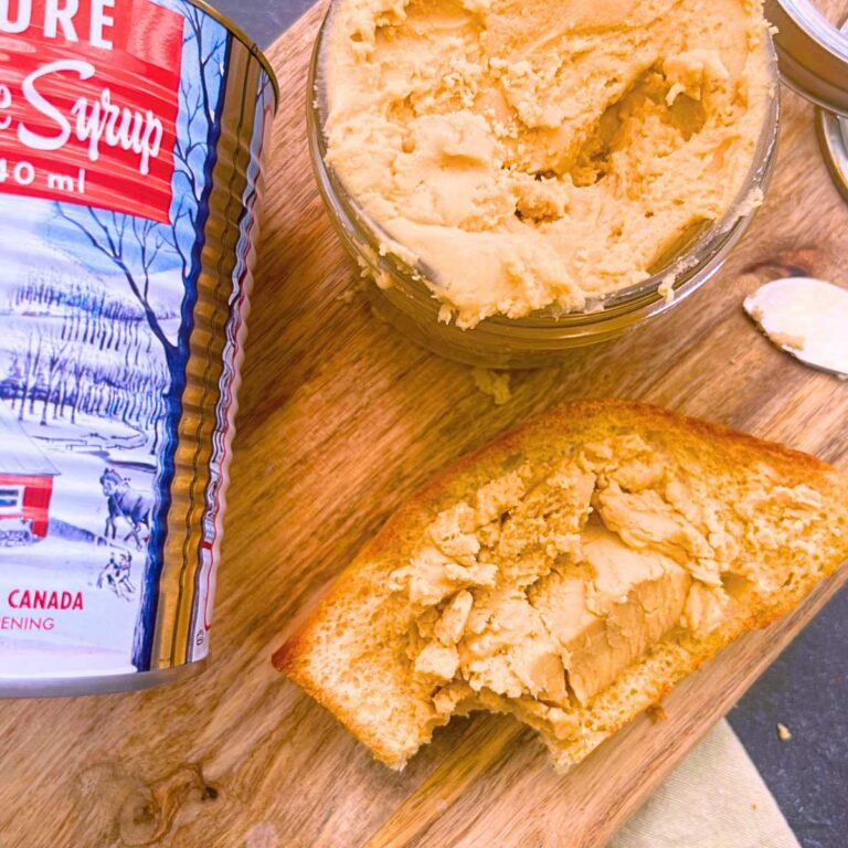 A wooden cutting board with a can of maple syrup, a glass bowl with maple butter, and a slice of toast with a bite out of it.