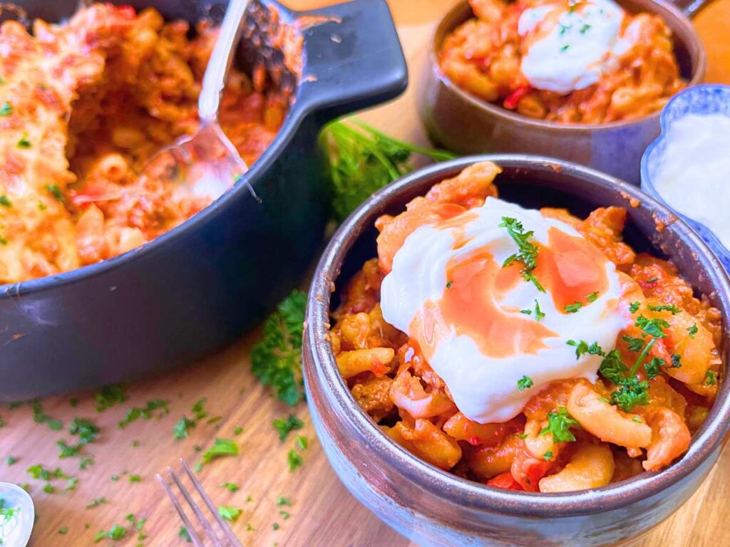 Two servings of chili mac in cheese in bowl topped with sour cream and hot sauce. There is more in a black casserole dish.