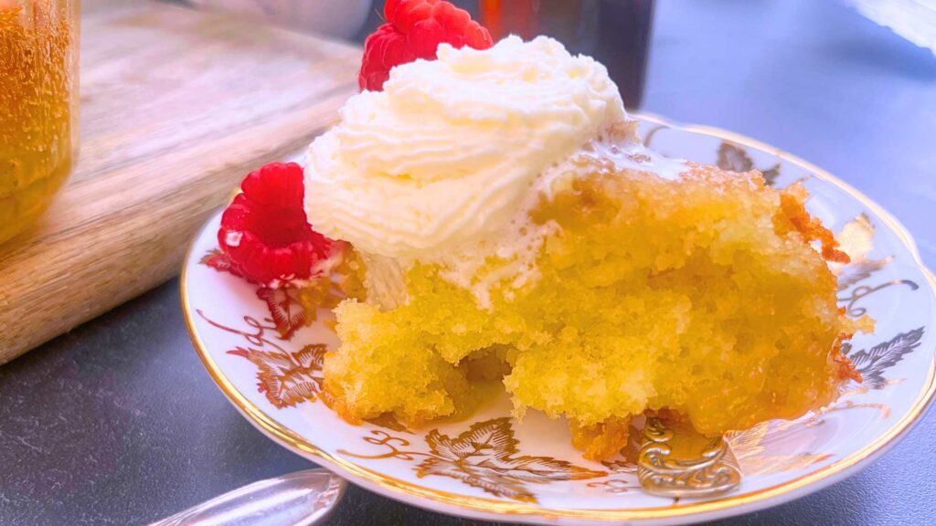 A serving of pouding chômeur on a white plate with gold maple leaves. There is whipped cream and raspberries on top.