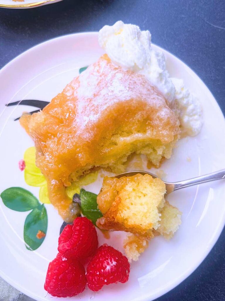 A serving of pouding chômeur on a white plate. There is a spoon and on the plate and the pouding is garnished with whipped cream and raspberries.