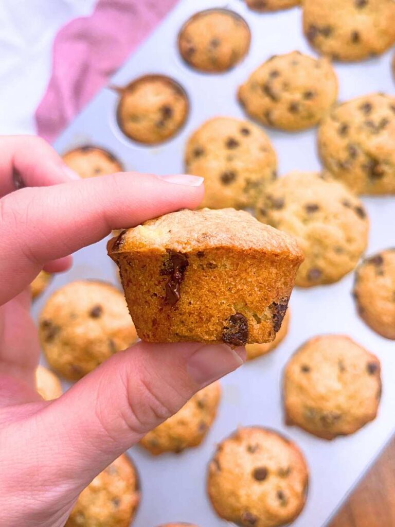 A woman is holding a mini banana chocolate chip muffin. There are more in a pan in the background.