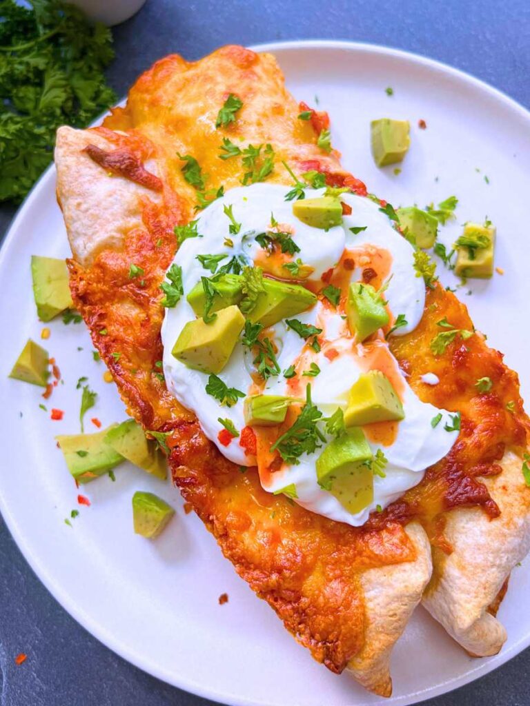 Two chicken enchiladas on a white plate. They are topped with sour cream, avacado, and hot sauce.