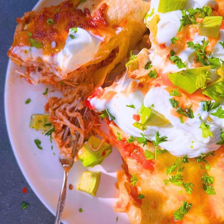An overhead view of chicken enchiladas on a white plate. They are topped with sour cream, avacado, and hot sauce.