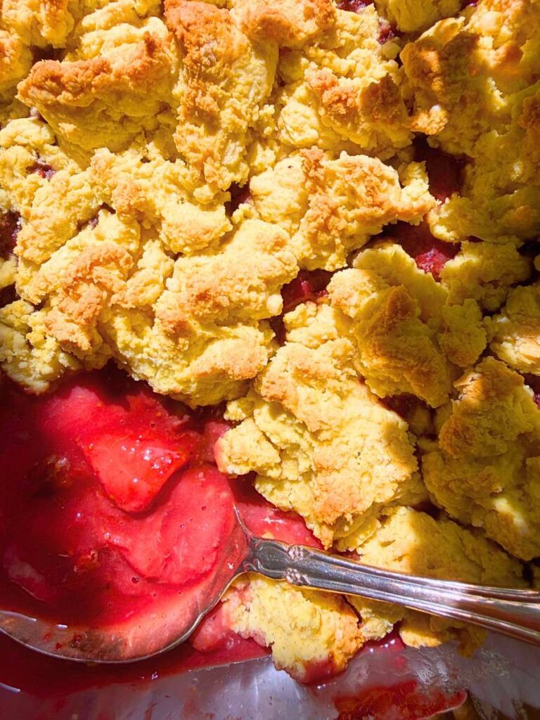 An overhead view of a baked strawberry cobbler with a scoop removed. There is a metal serving spoon inside.