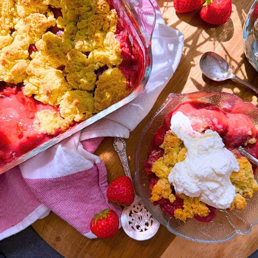 A baked strawberry dessert with a biscuit topping. There is a serving in front in a glass bowl. There is whipped cream on top.